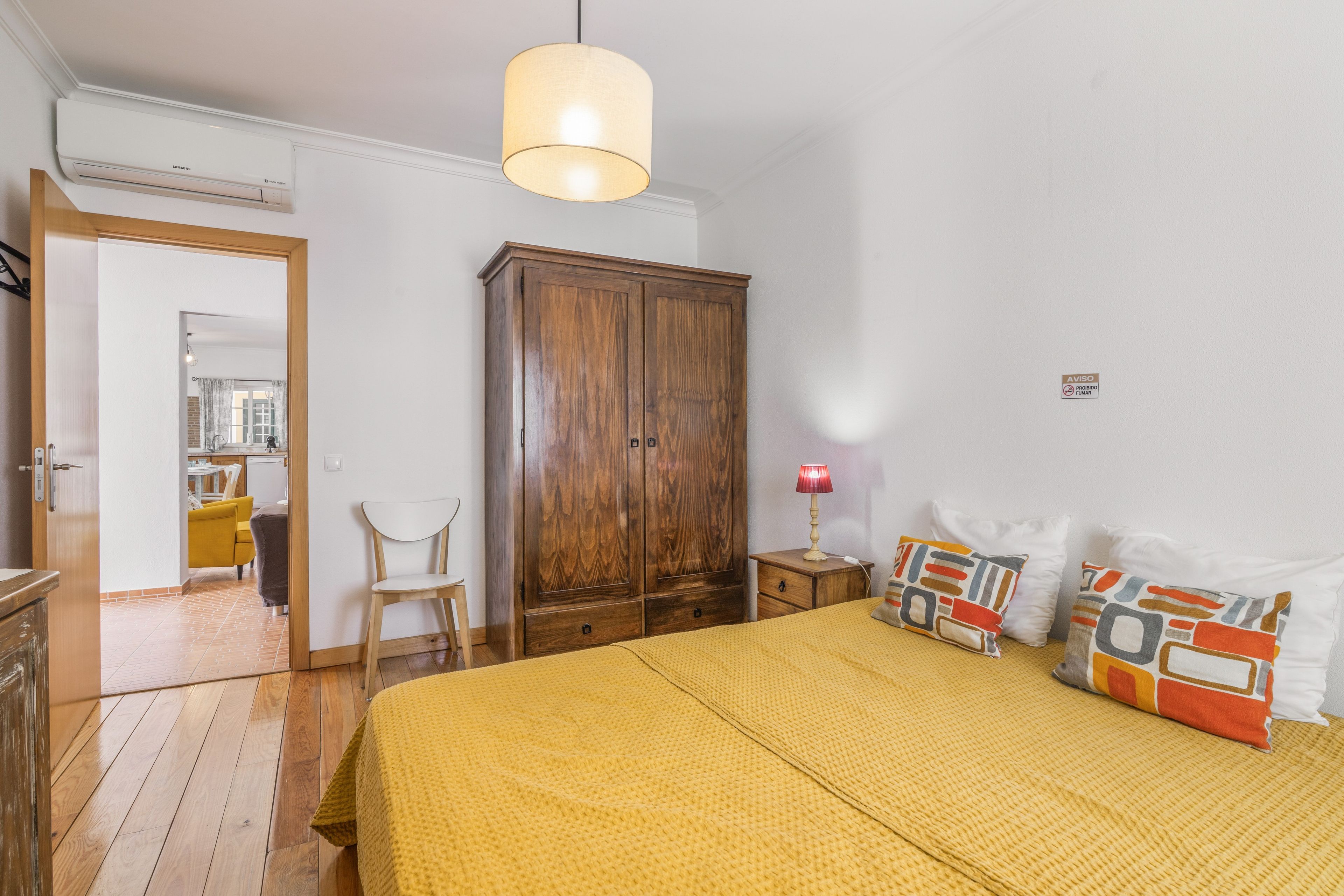 Ein Schlafzimmer mit einem Holzschrank, einem Bett mit gelber Bettwäsche, einer roten Lampe und Blick in den angrenzenden Raum.