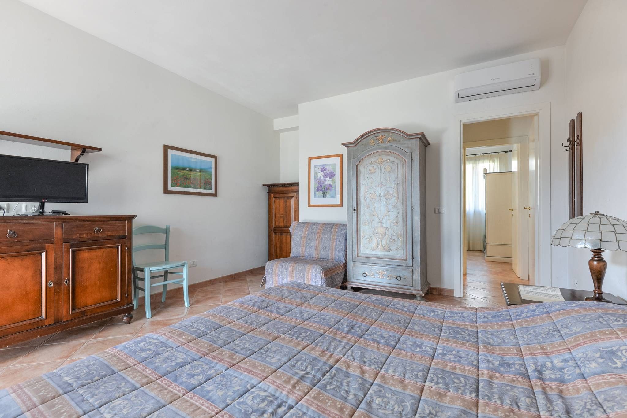 A bedroom with a bed, a wooden dresser, a television, a cabinet, and a door leading to another room.