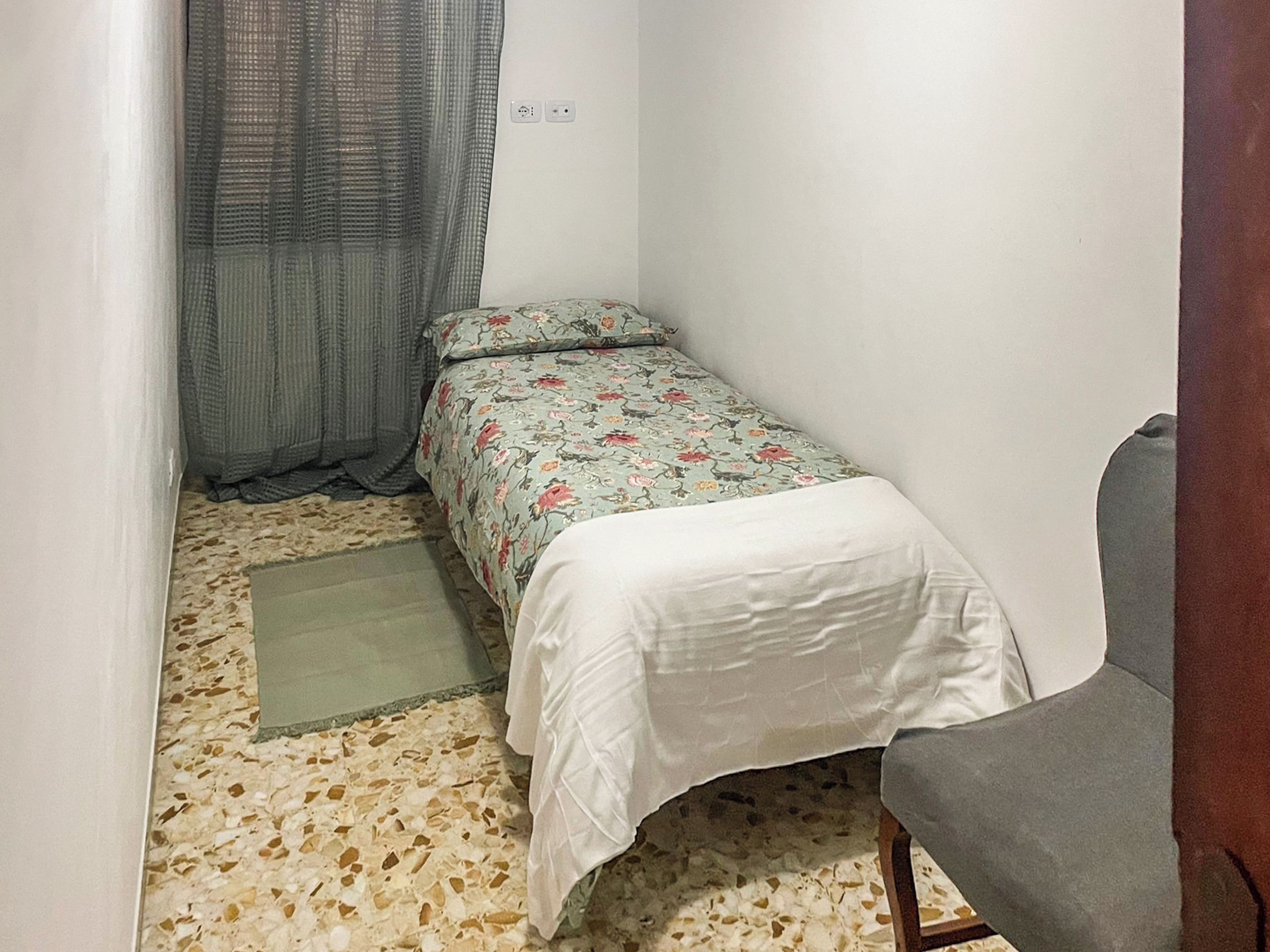 Une petite chambre avec un lit simple, une couvre-lit à motifs floraux, une chaise grise et un sol à motifs.