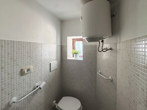 A small bathroom with a toilet, a handrail, a towel rack, and a window with a plant. - I Rifugi dei Lucretili (Licenza)