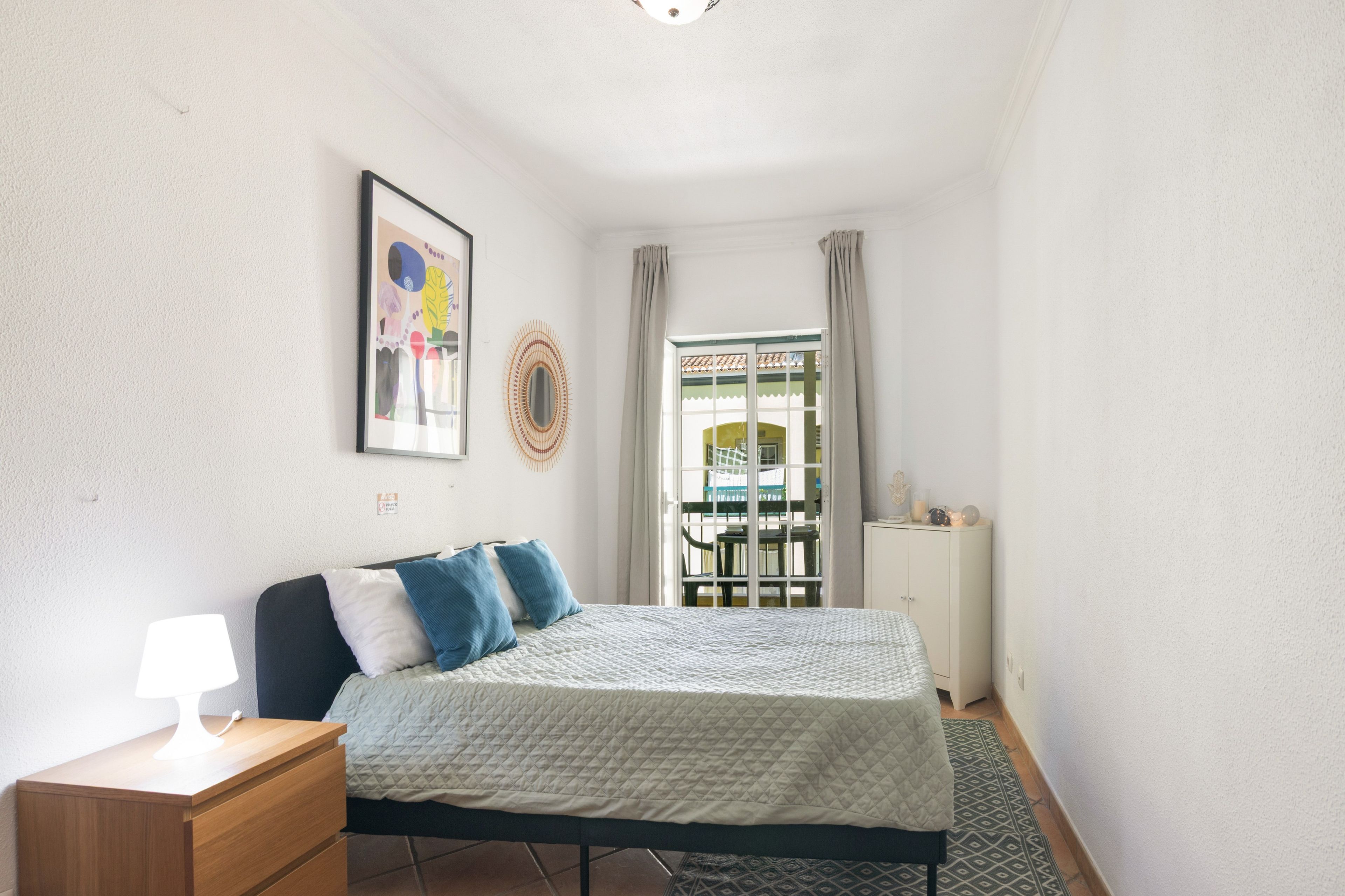 Ein Schlafzimmer mit einem Bett, einem Nachttisch mit Lampe, einem Wandbild, einem Fenster mit Vorhängen und Blick ins Freie.