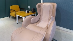Una silla reclinable con un respaldo alto y reposabrazos, una mesa de madera con una taza blanca y un objeto negro, y una silla amarilla al fondo.