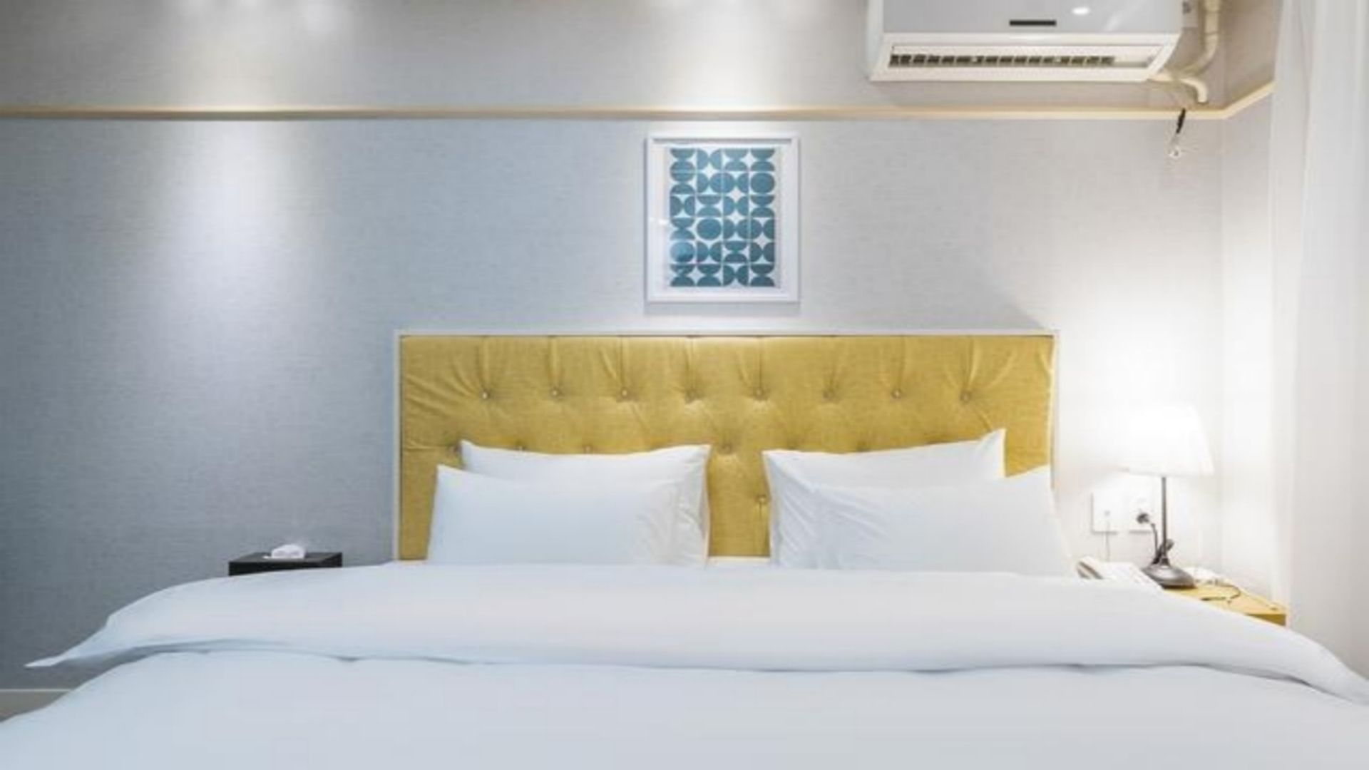 Hotellihuone, jossa on sänky, yöpöytä, lamppu ja seinälle asennettu ilmastointilaite.