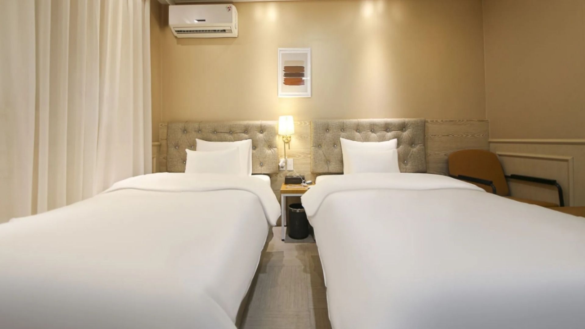 A hotel room with two beds, a bedside table with a lamp, an air conditioning unit, and a framed picture on the wall.