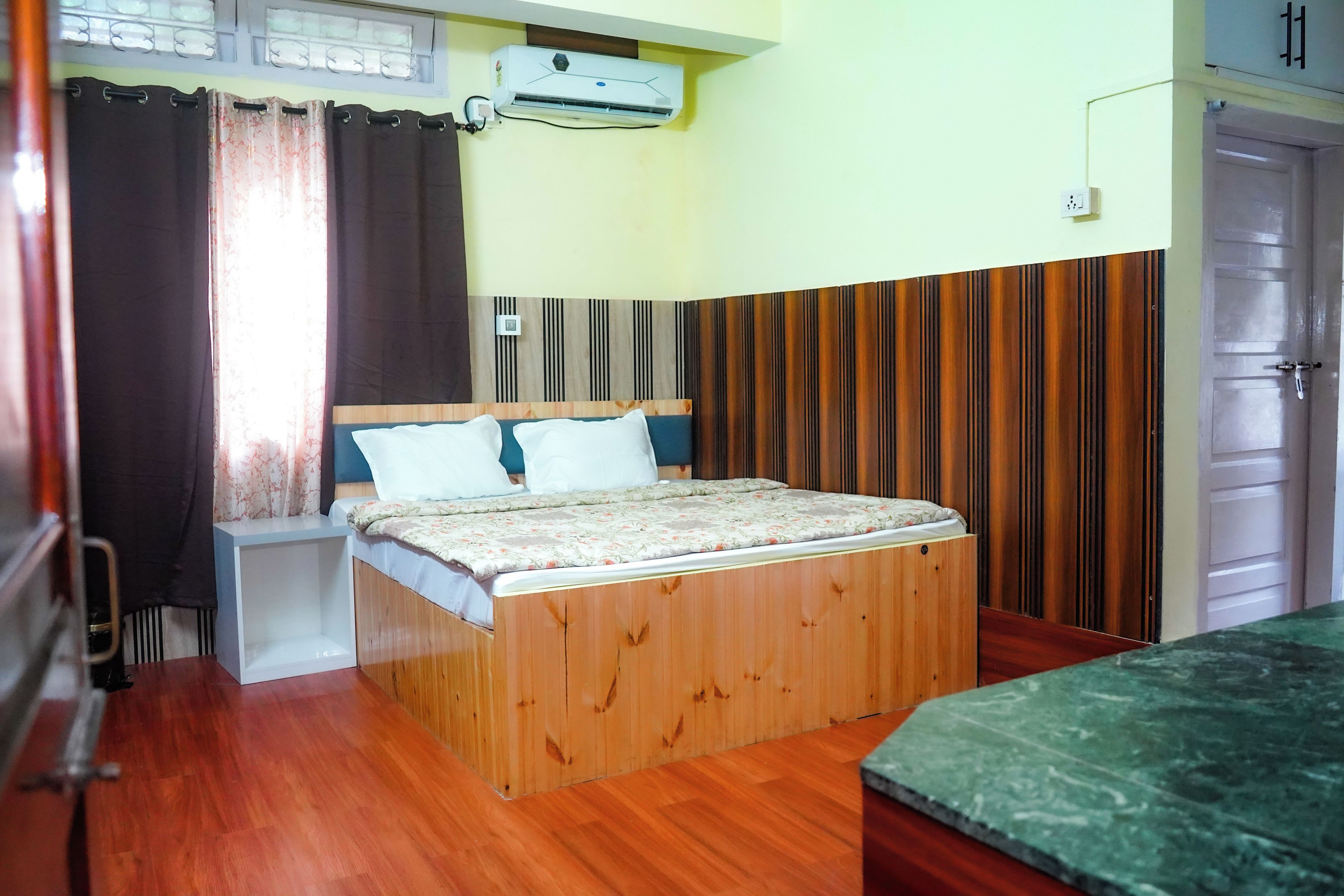 A room with a wooden bed, a small bedside table, a wall-mounted air conditioner, a window with curtains, and a door.