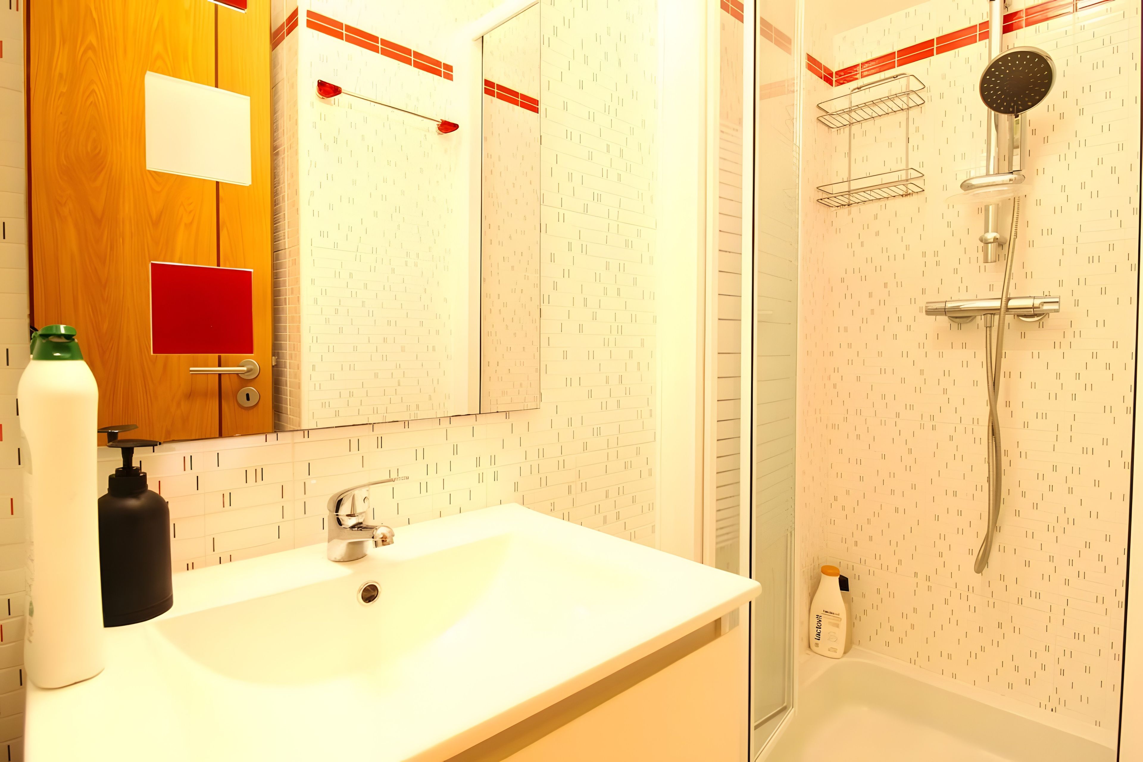 Un baño con un lavamanos blanco, un espejo, una ducha con cabezal de ducha manual y una botella de loción.
