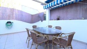 Um pátio externo com uma mesa redonda e cadeiras de vime, uma área coberta com toldo listrado e uma mangueira de metal enrolada no chão.