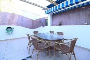 Un patio exterior con una mesa redonda y sillas de mimbre, un espacio cubierto con toldo a rayas y una manguera metálica enrollada en el suelo.
