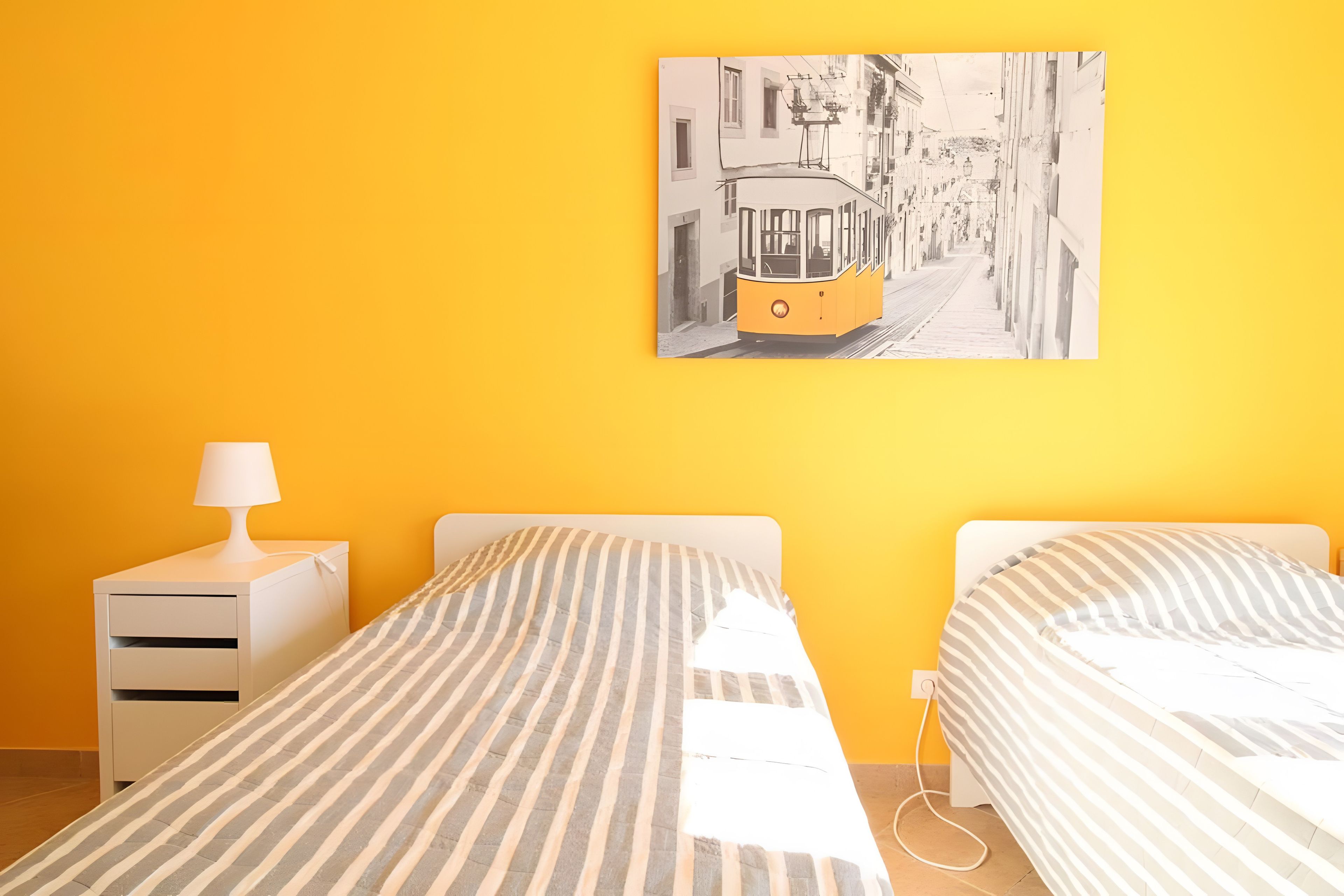 Una habitación con dos camas, una mesita de noche, una lámpara y un cuadro en la pared que muestra un tranvía.