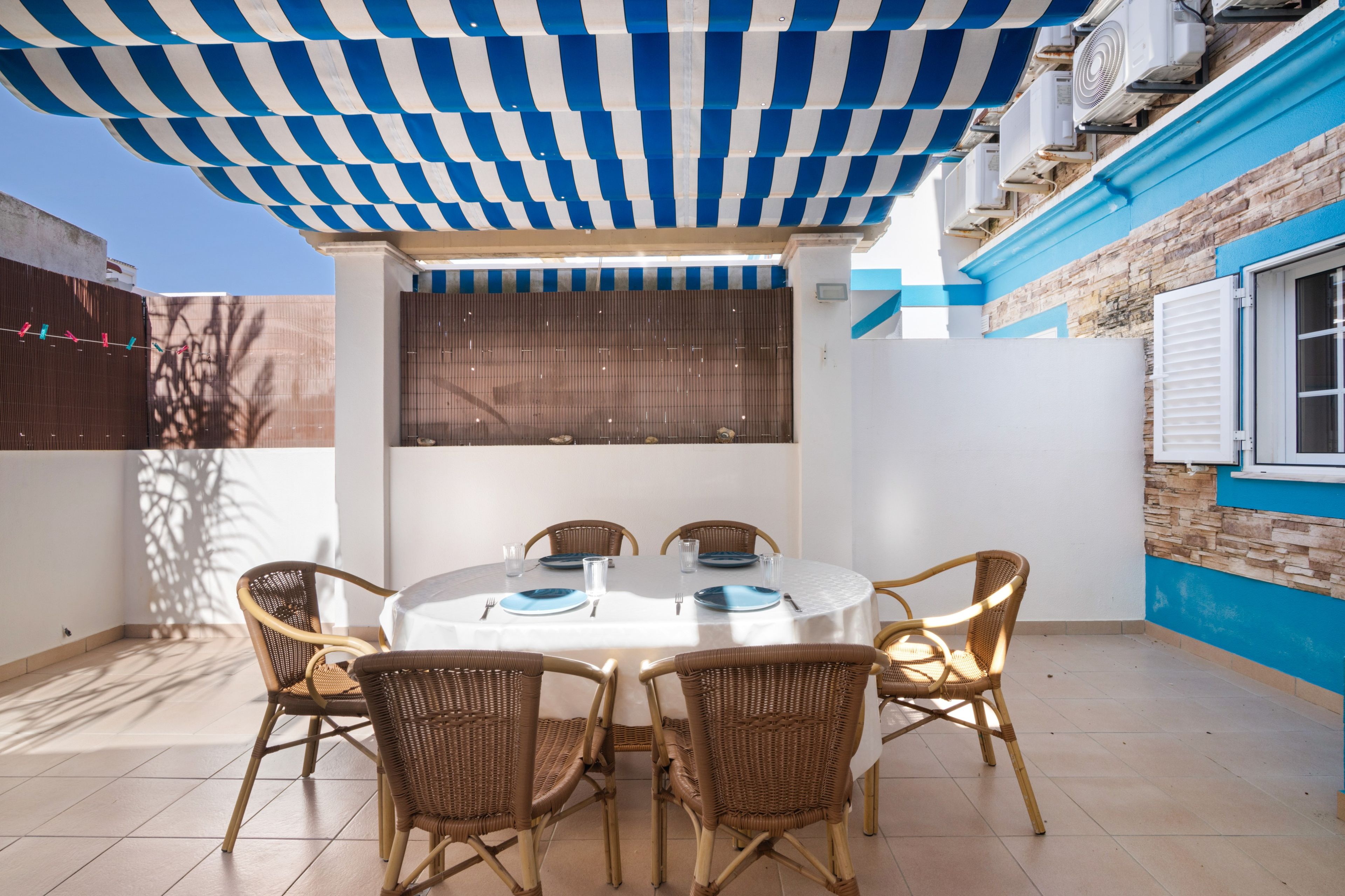 A covered outdoor dining area with a table and chairs, a window with blinds, and a wall with a blue and white pattern.