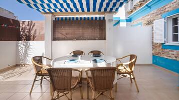 A covered outdoor dining area with a table and chairs, a window with blinds, and a wall with a blue and white pattern.