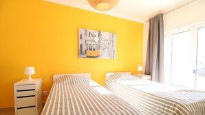 Quarto de hotel com duas camas, uma mesa de cabeceira, uma luminária, um quadro na parede e vista para o exterior.