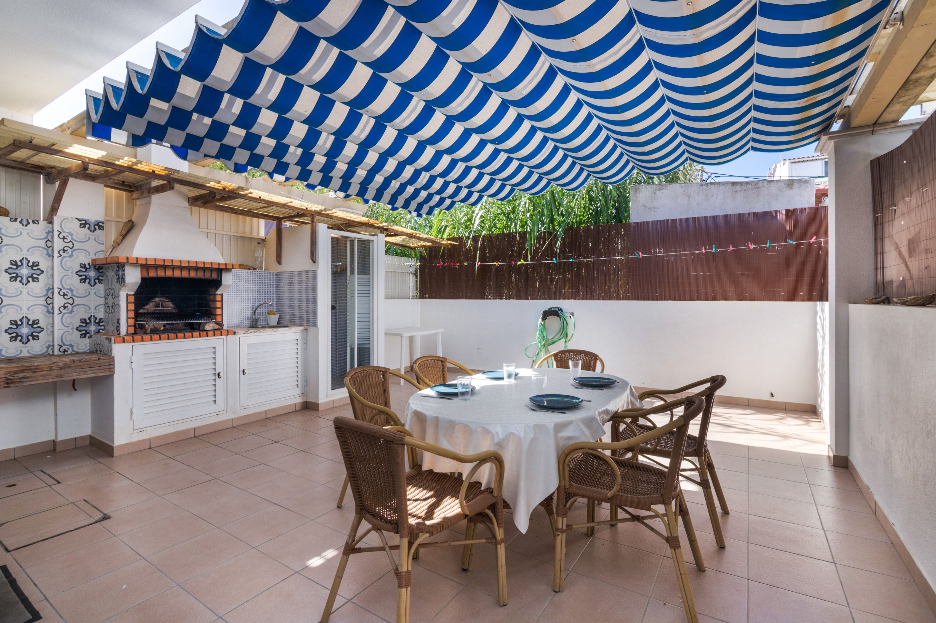 Un patio exterior con una mesa redonda y sillas de mimbre, una parrilla y un toldo a rayas azules.