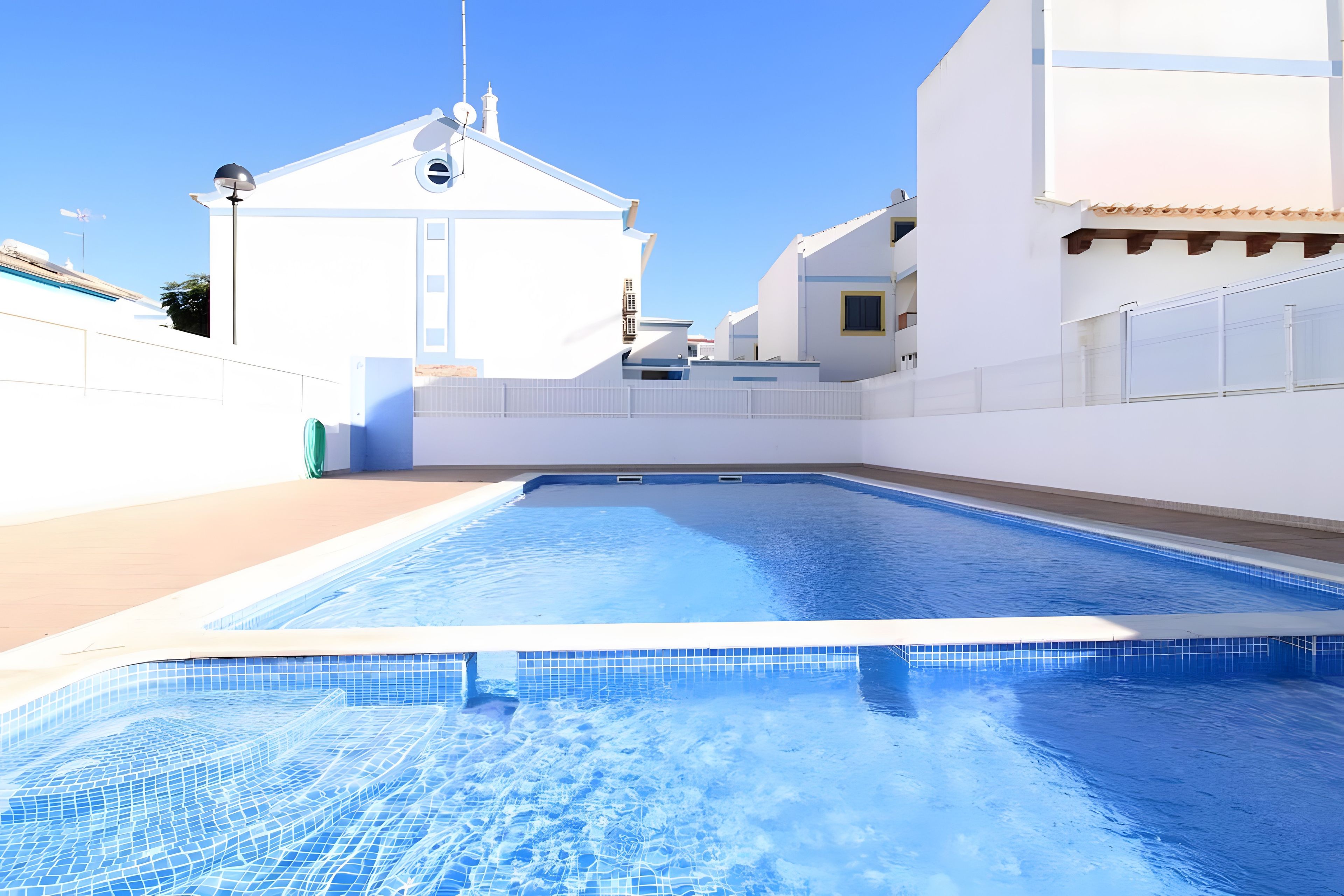 Una piscina con borde de azulejos, rodeada de edificios blancos y un cielo despejado.