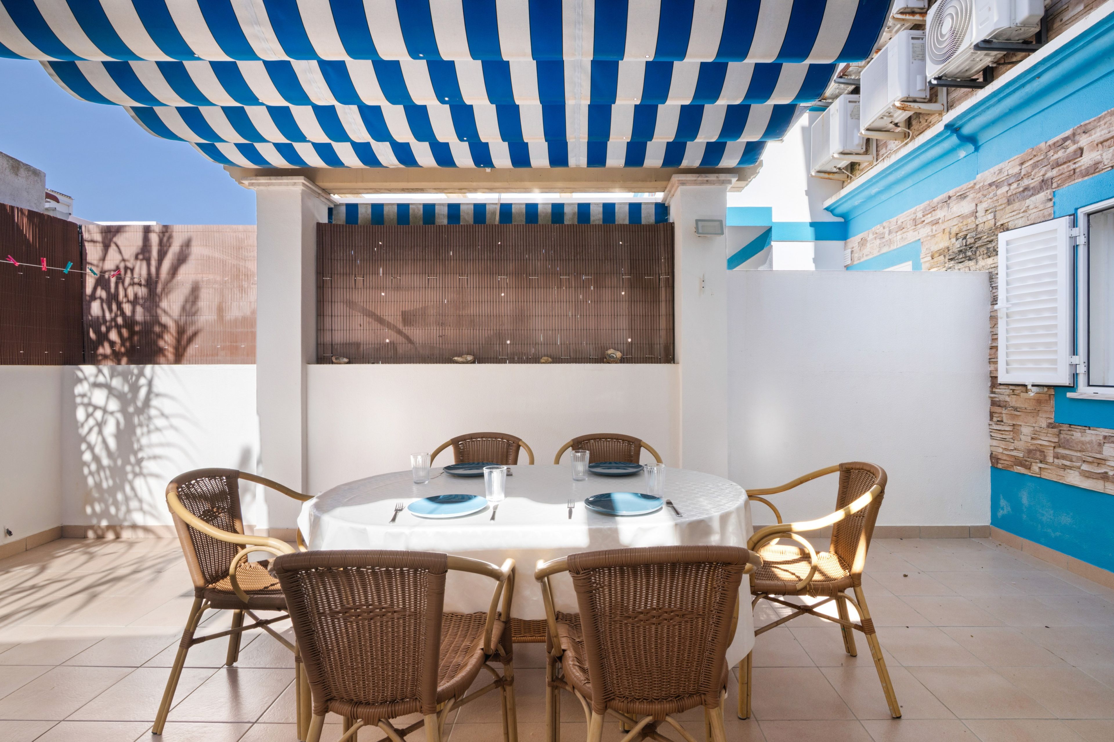 Un espace extérieur abrité, aménagé pour les repas, avec une table ronde, six chaises en osier et un auvent rayé de bleu et de blanc.