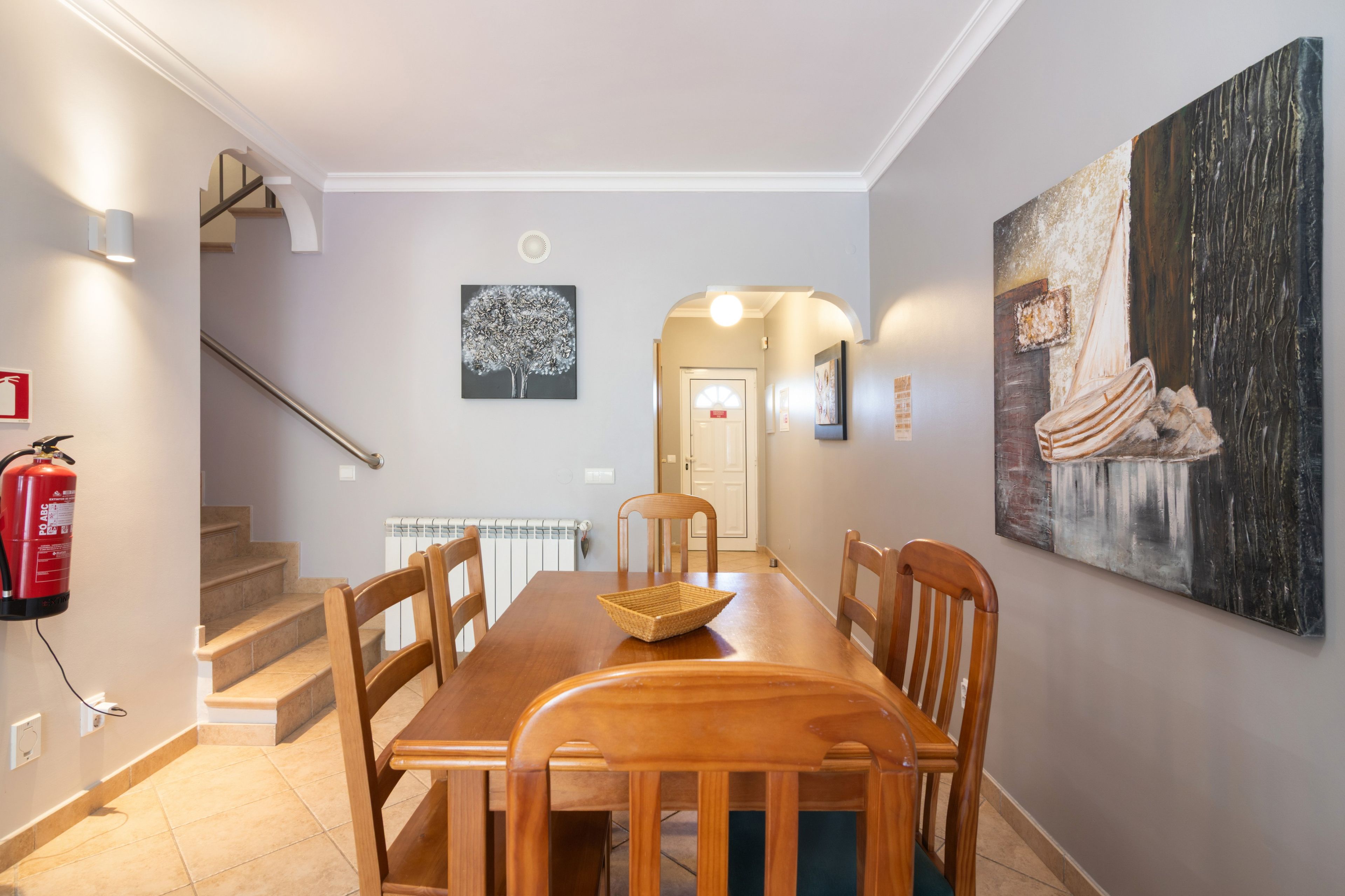 Un comedor con una mesa y sillas de madera, un extintor, una escalera y un pasillo que conduce a una puerta.