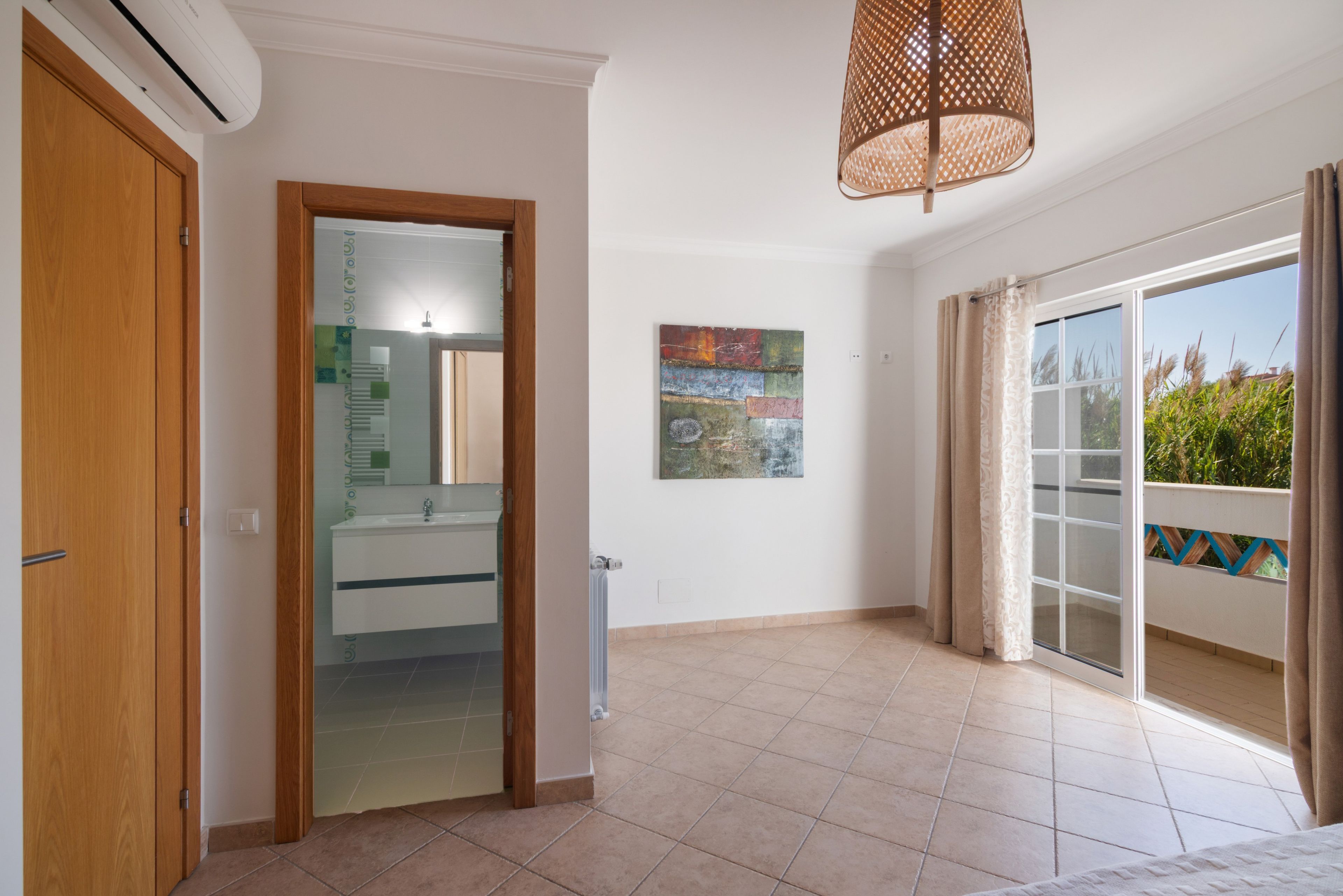 Une pièce dotée d’une porte en bois, d’une porte en verre donnant sur une salle de bain, d’un ventilateur de plafond, d’une œuvre d’art accrochée au mur et d’une vue sur l’extérieur par une fenêtre avec des rideaux.