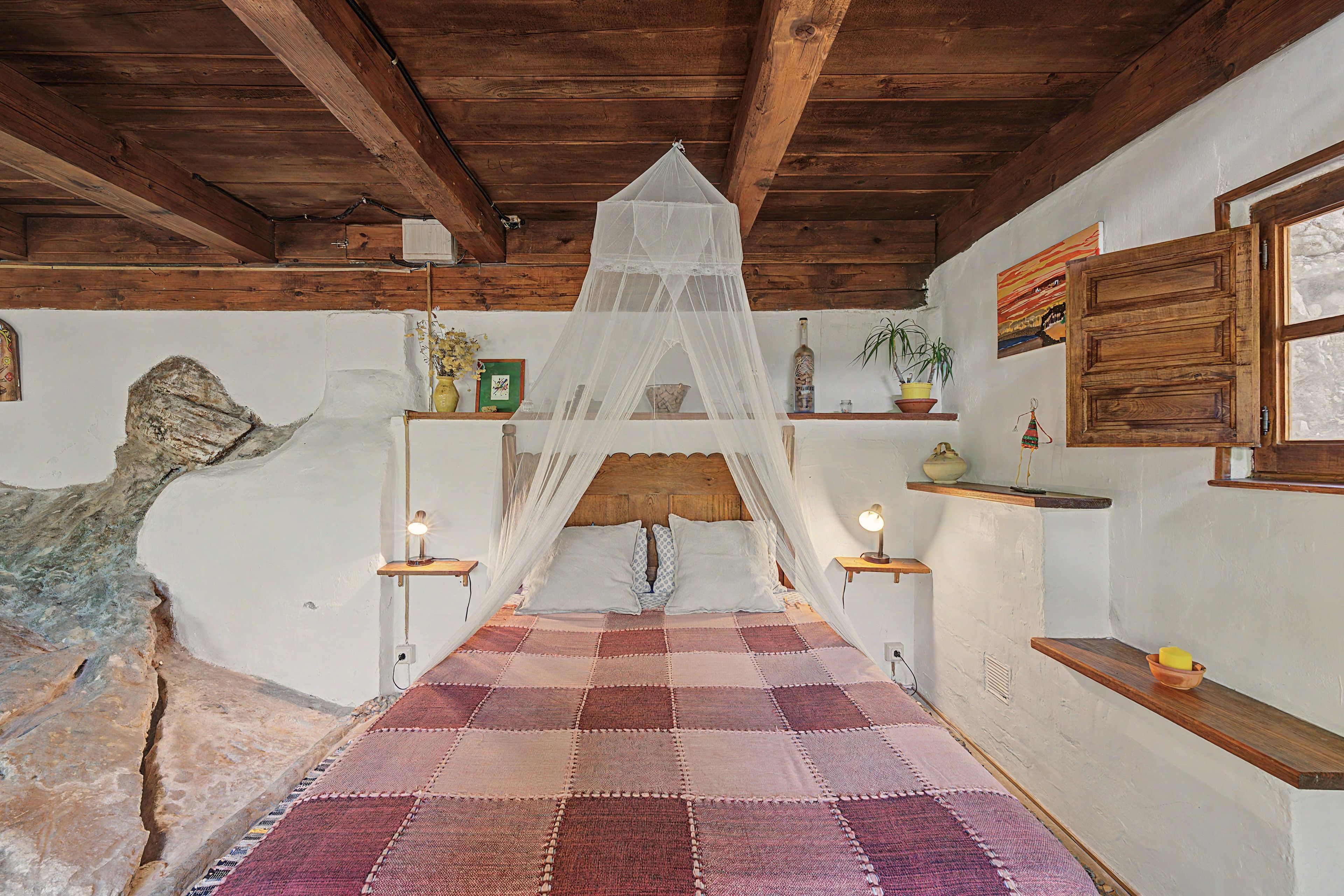 A room with a checkered floor, wooden beams, a bed with a canopy, bedside lamps, and a rustic wall.