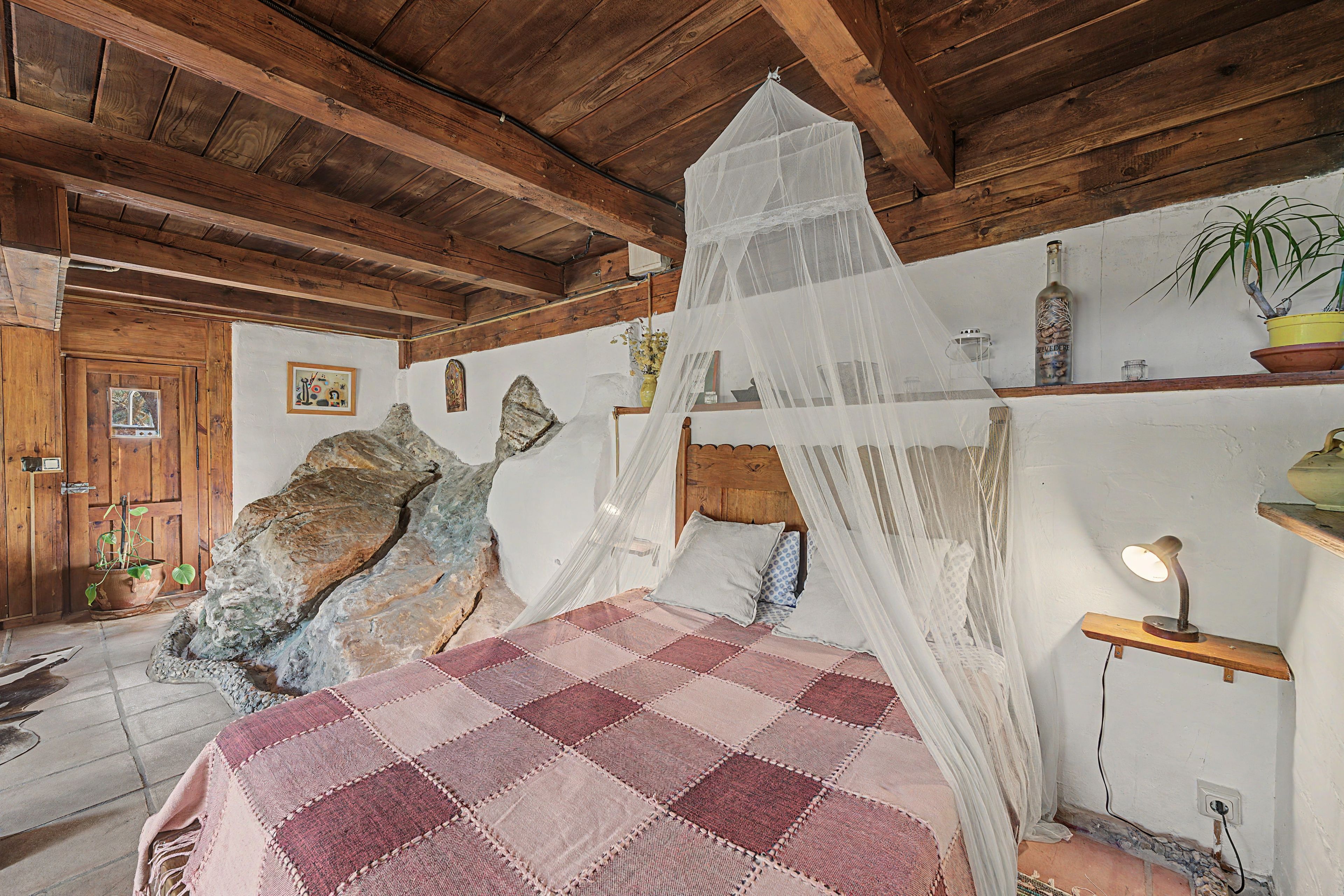 Un dormitorio rústico con una cama bajo dosel, vigas de madera en el techo, una pared de piedra y un estante con plantas y una botella.