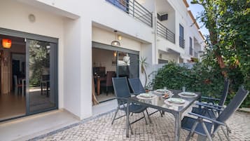 Un área de comedor exterior moderna con una mesa y sillas de vidrio, una planta en maceta y vistas del interior del edificio.