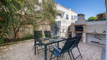 Un patio exterior con mesa lista para comer, rodeado de sillas y una zona de barbacoa.