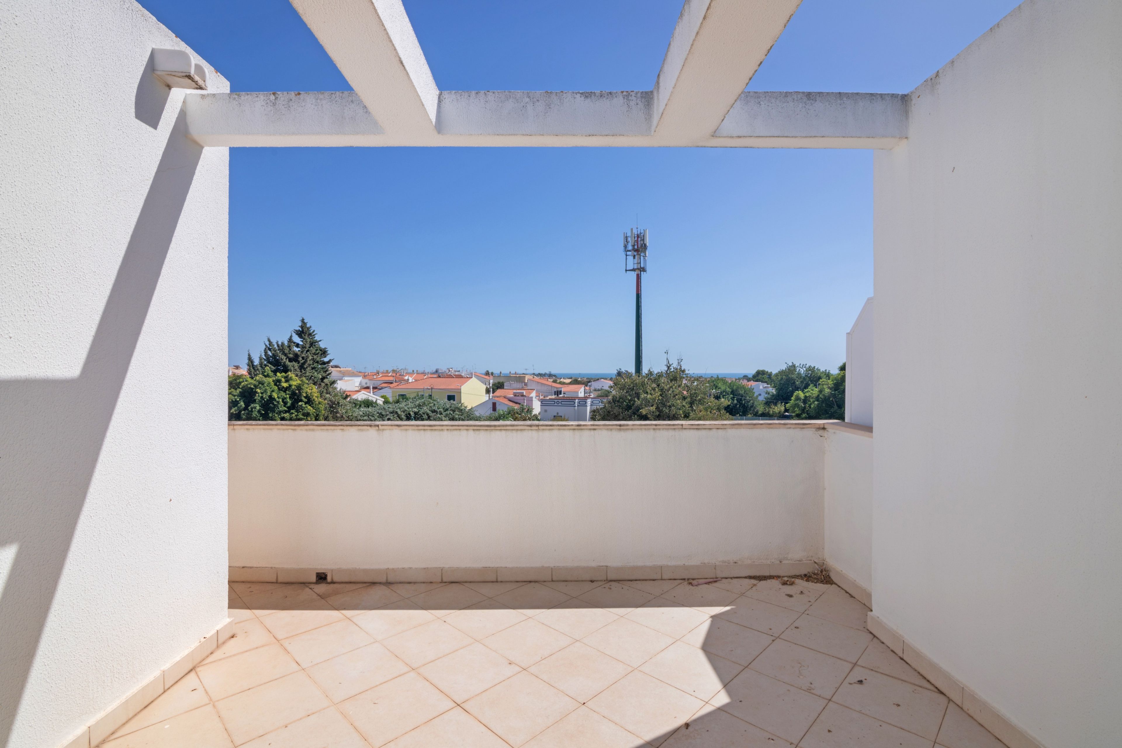 A balcony with a tiled floor and white walls, overlooking a residential area with houses and a communication tower.