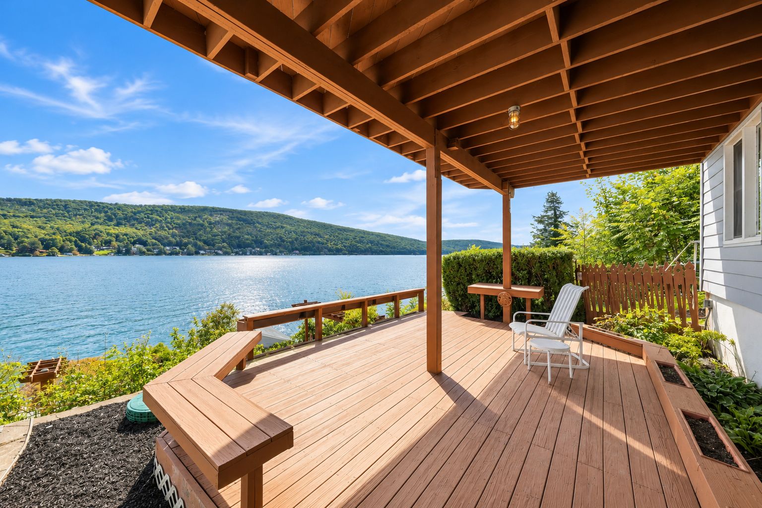 A wooden deck with a pergola, a table, and two chairs overlooking a lake with a forested hill in the background.