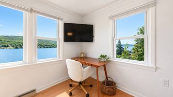 Une pièce comprenant un bureau, une chaise et un téléviseur fixé au mur. Deux fenêtres offrent une vue sur un lac et la verdure environnante.