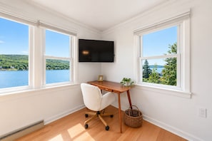 A room with a desk, chair, and a TV mounted on the wall. There are two windows with a view of a lake and greenery.