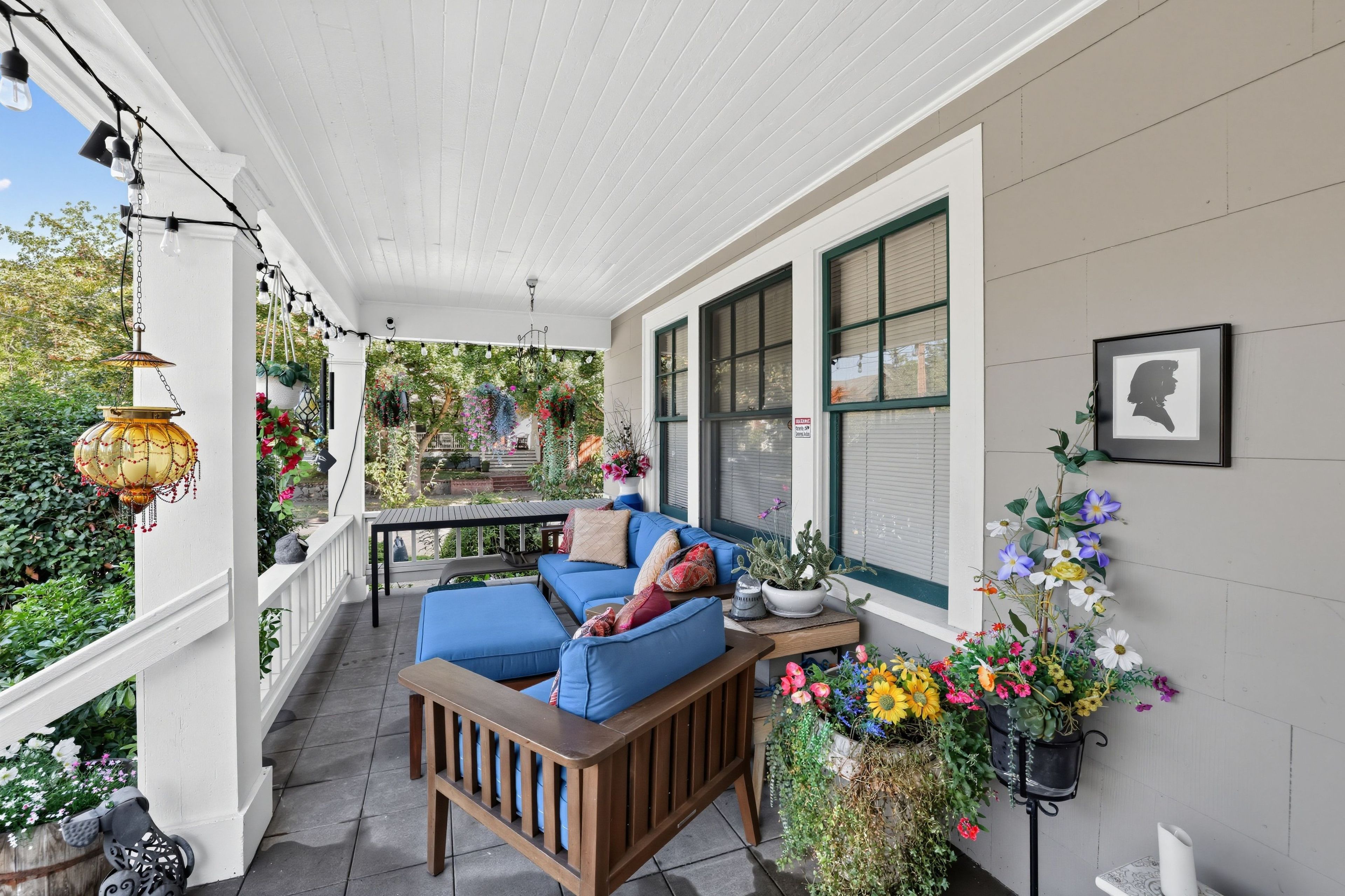 A covered porch with a wooden bench and cushions, potted plants, and hanging lights.