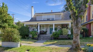 Une maison à deux étages avec un porche à l’avant, une cheminée et un petit jardin devant, avec un arbre et un banc.