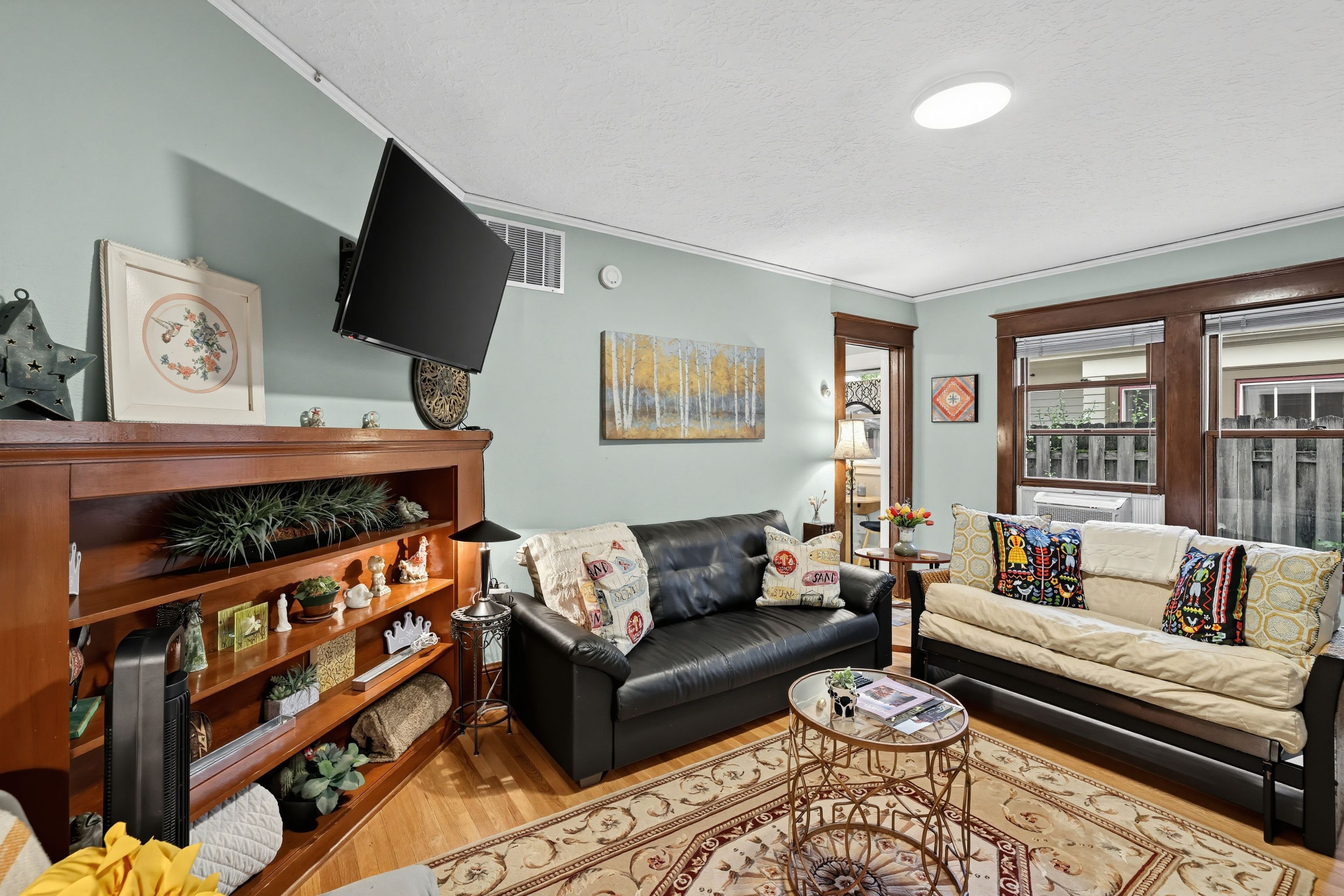 A living room with a black leather sofa, a beige sofa, a wooden bookshelf, a flat-screen TV, and a patterned rug.