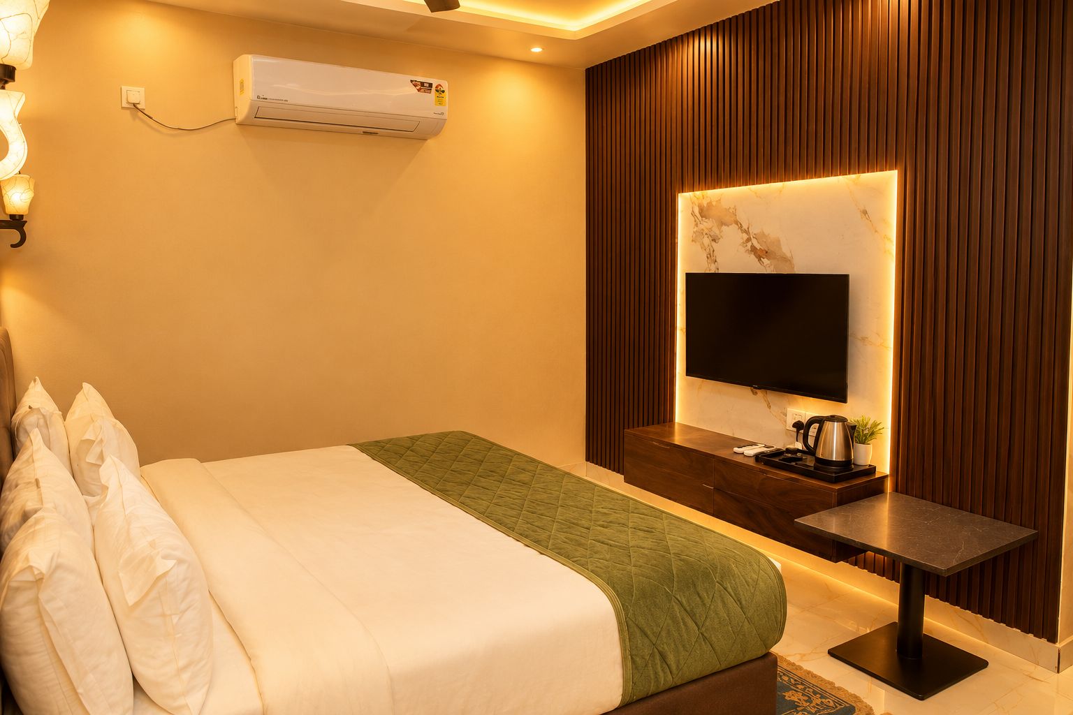 A hotel room with a bed, a flat-screen TV mounted on a wall, a bedside table with a coffee maker, and an air conditioning unit on the wall.