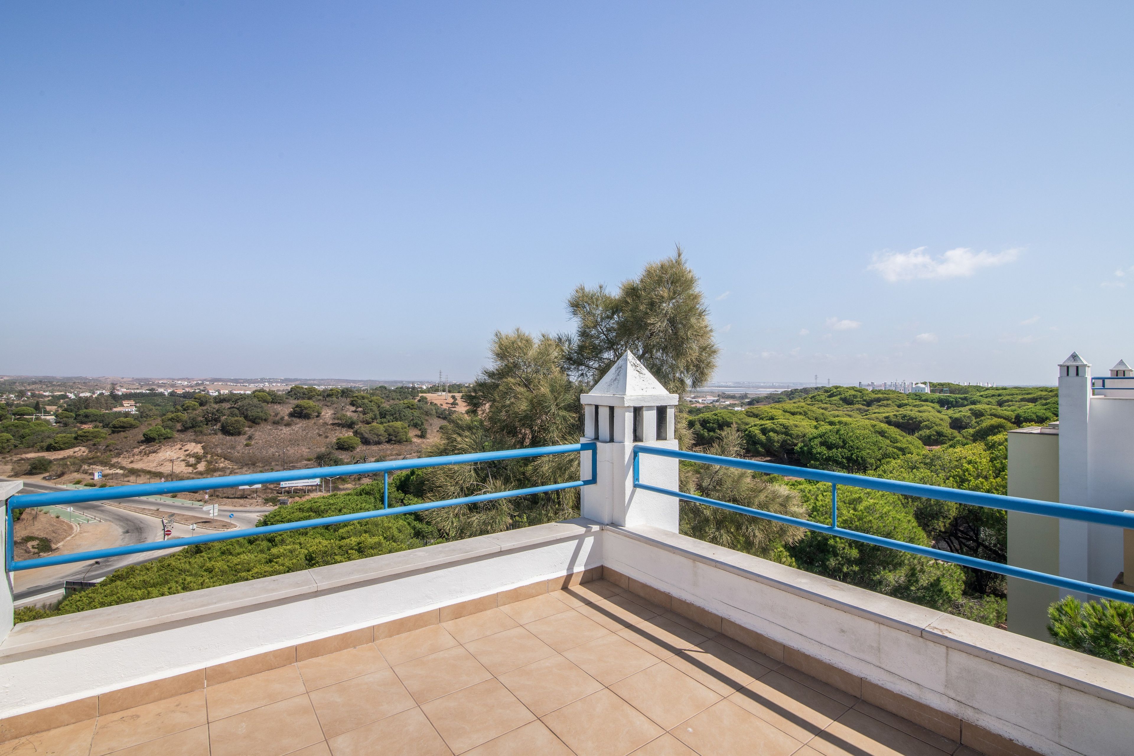 Una terraza con vista a un paisaje urbano, una estructura blanca con techo a dos aguas y una barandilla azul.