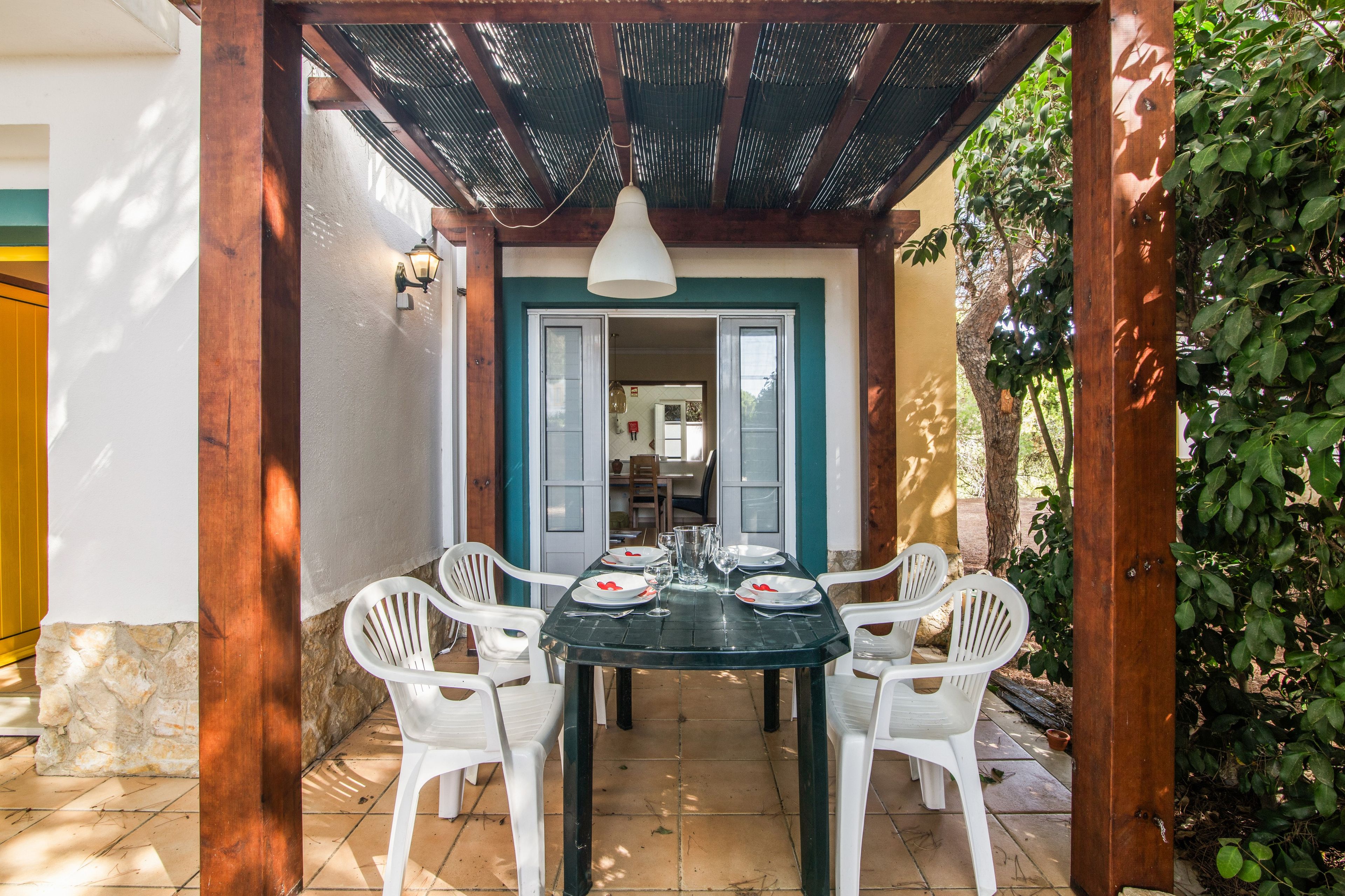 Een terras buiten met een tafel voor vier personen, omringd door witte stoelen, onder een houten pergola.