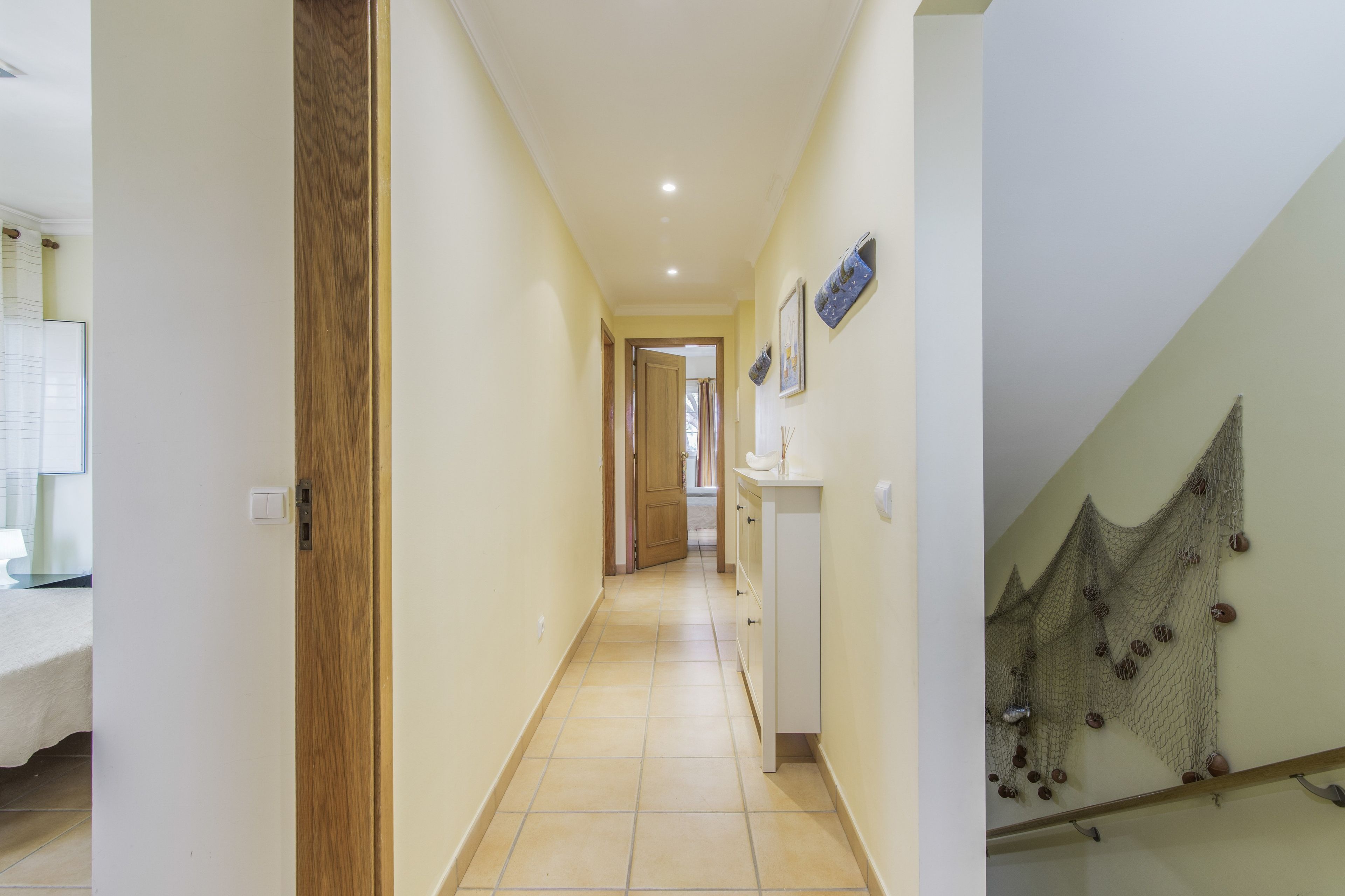 Un couloir avec une porte en bois, un mur blanc, un sol carrelé et un escalier orné d’un motif en réseau.
