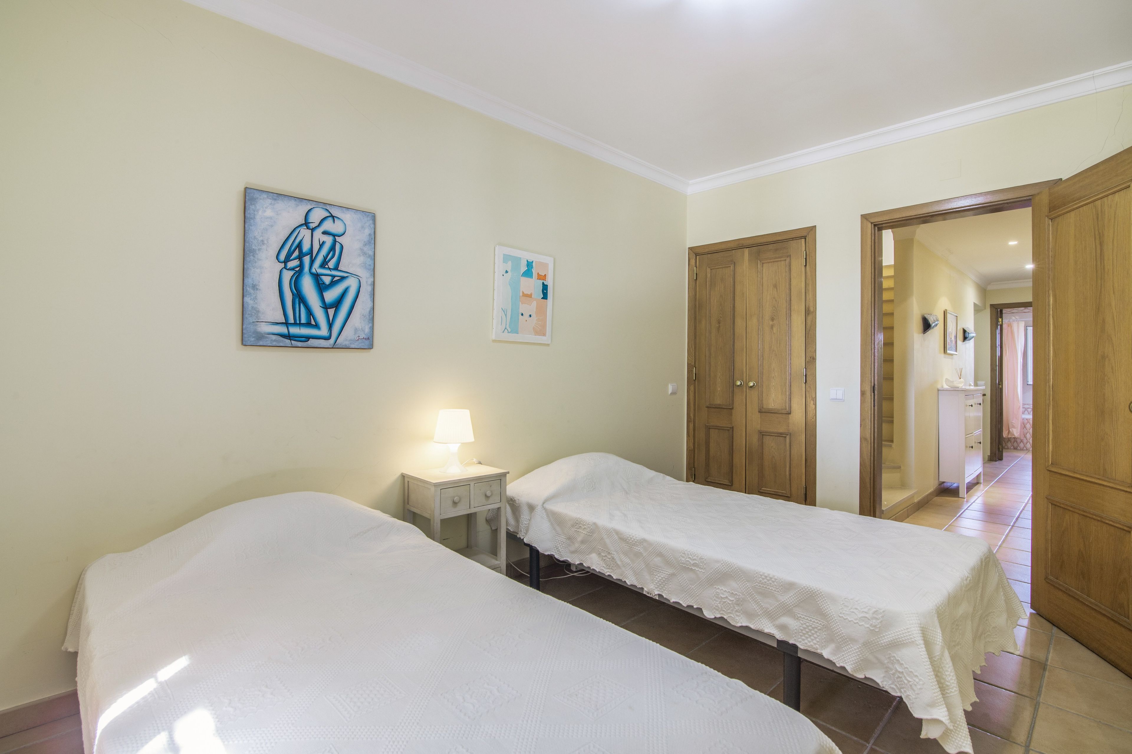 Habitación de hotel con dos camas individuales, una mesita de noche con lámpara, un cuadro en la pared y un armario de madera.