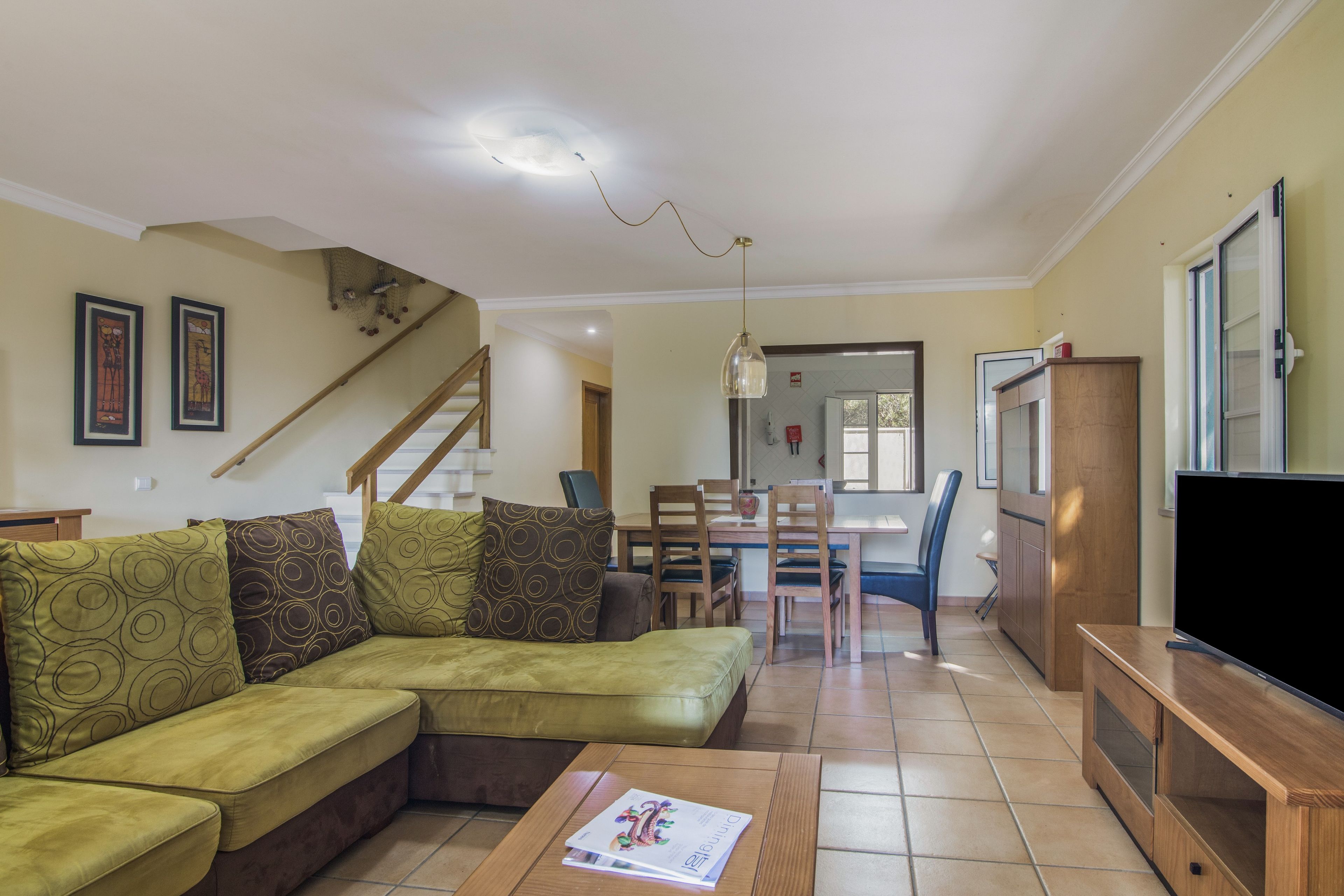A living room with a sectional sofa, a dining table with chairs, a television, and a staircase leading to another level.