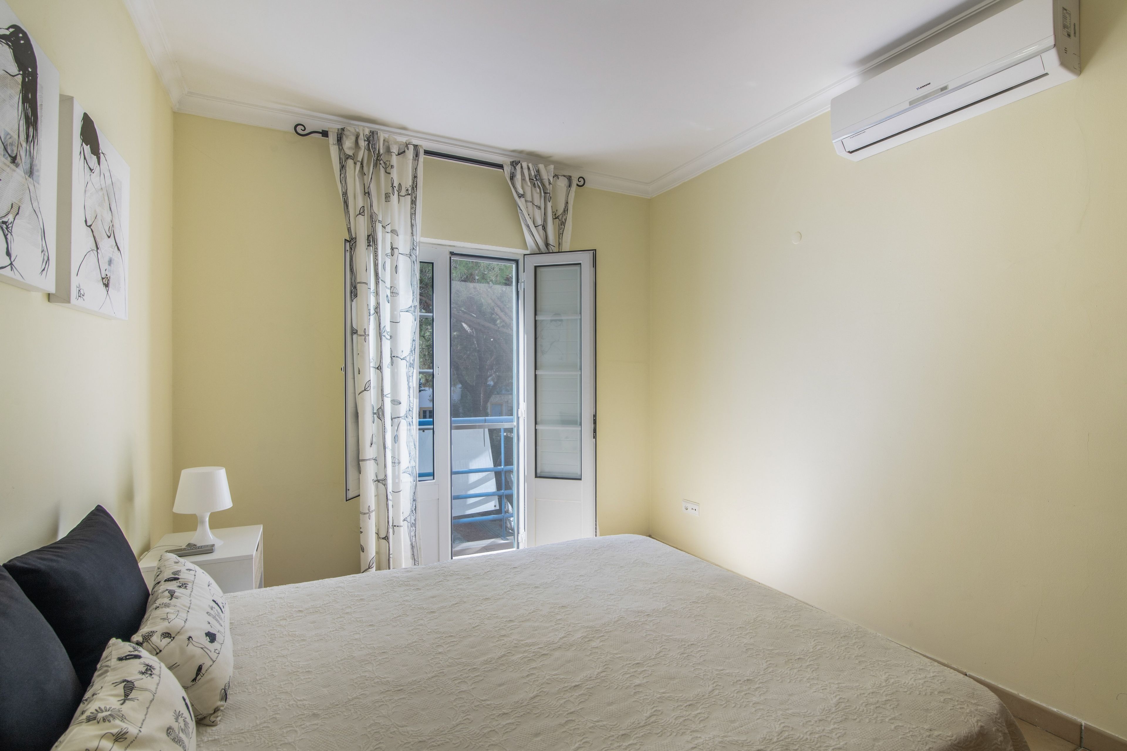 Un dormitorio con cama, mesita de noche, lámpara, ventana con cortinas y aire acondicionado.