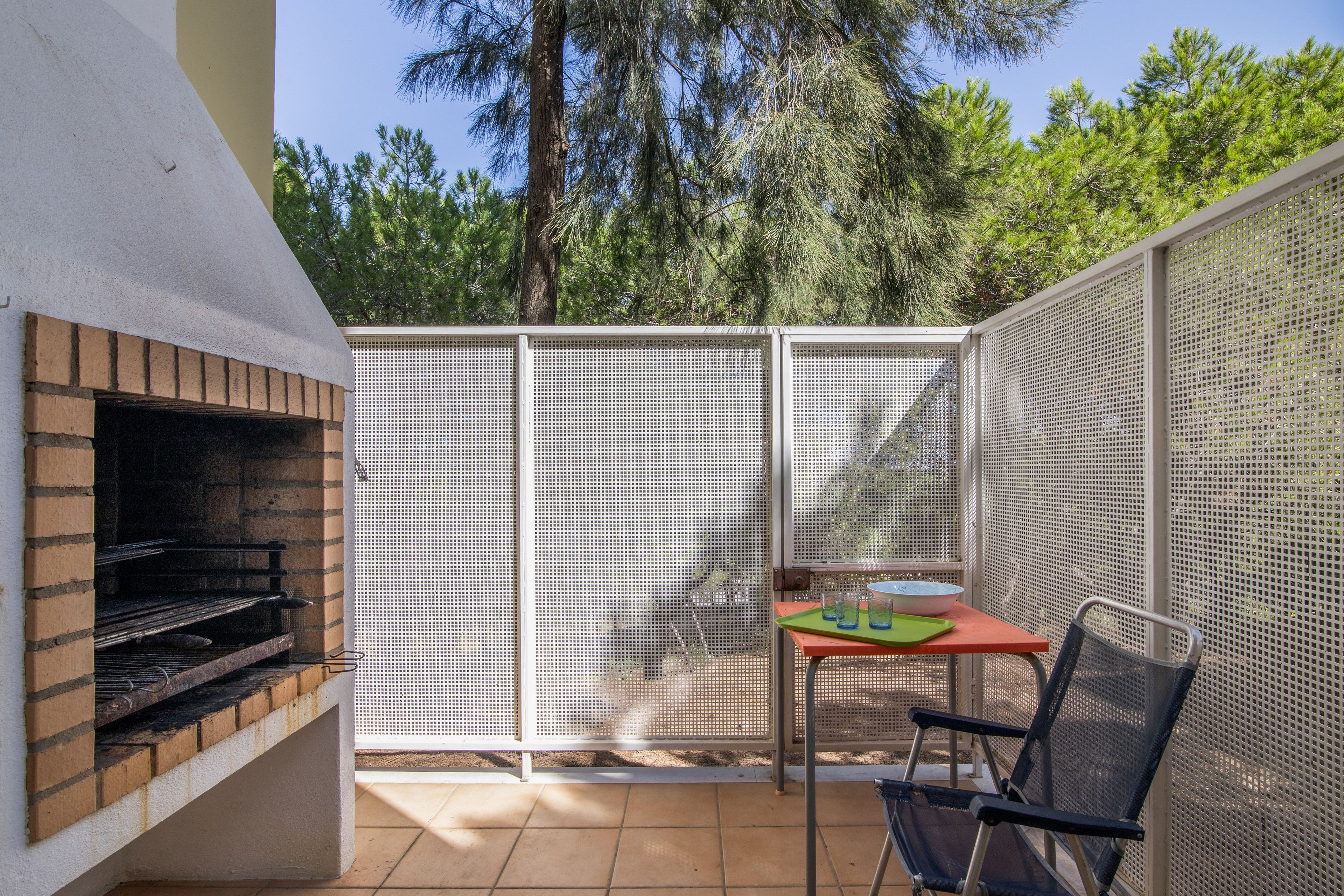 Een terras met een bakstenen barbecue, een tafel met een schaal en glazen, en een stoel.