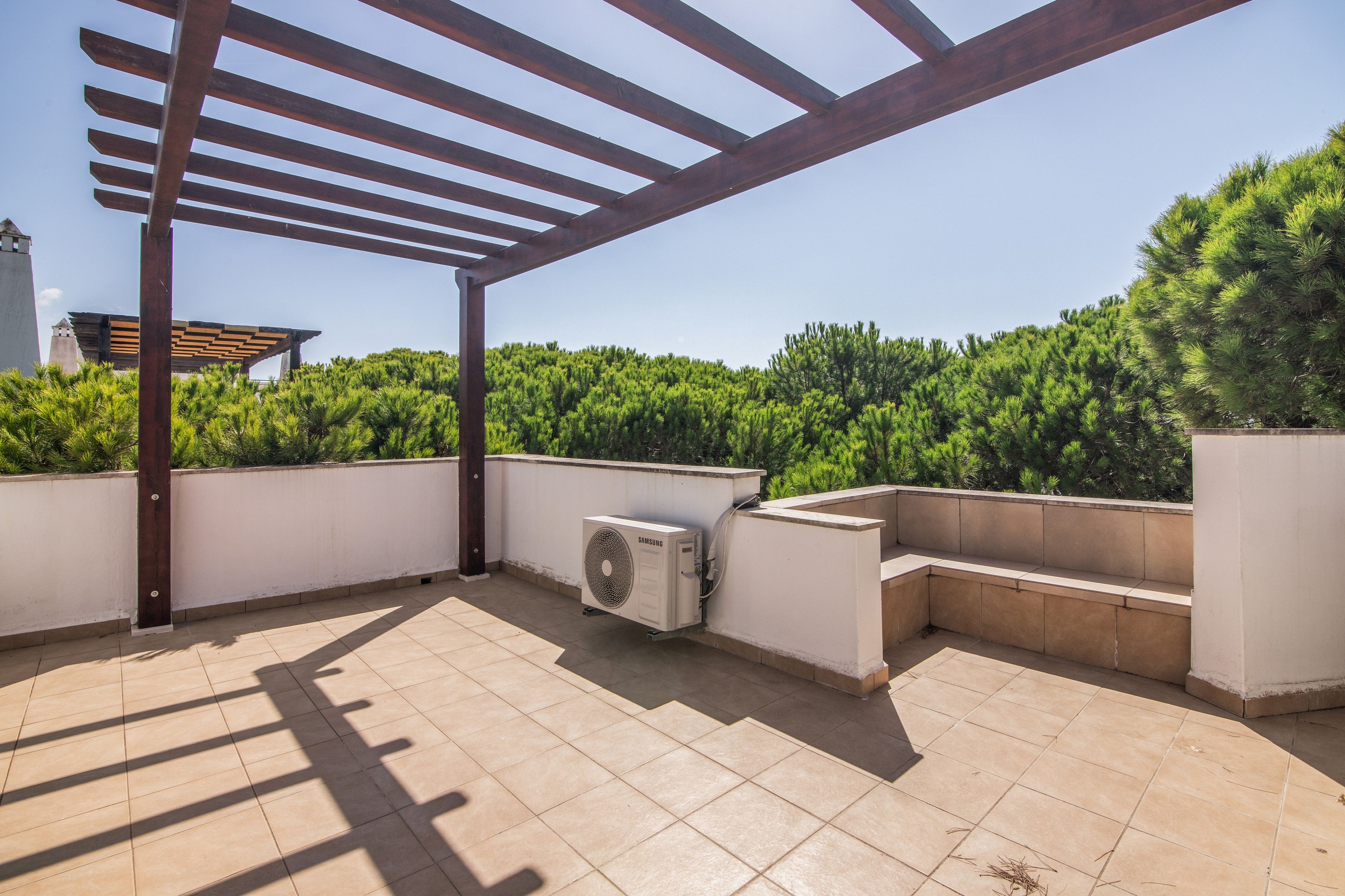 Un espace extérieur aménagé en patio, doté d’une pergola, d’un climatiseur Samsung et d’un banc.