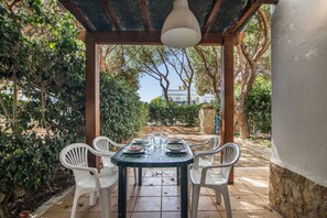 Un pati exterior amb taula preparada per a dinar, envoltat de cadires blanques, sota una pergola amb una làmpada penjant.
