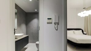 Et moderne badeværelse med en hvid vask, mørke skabe og et toilet. Der er et natbord med en lampe og en seng med hvide sengetøj.