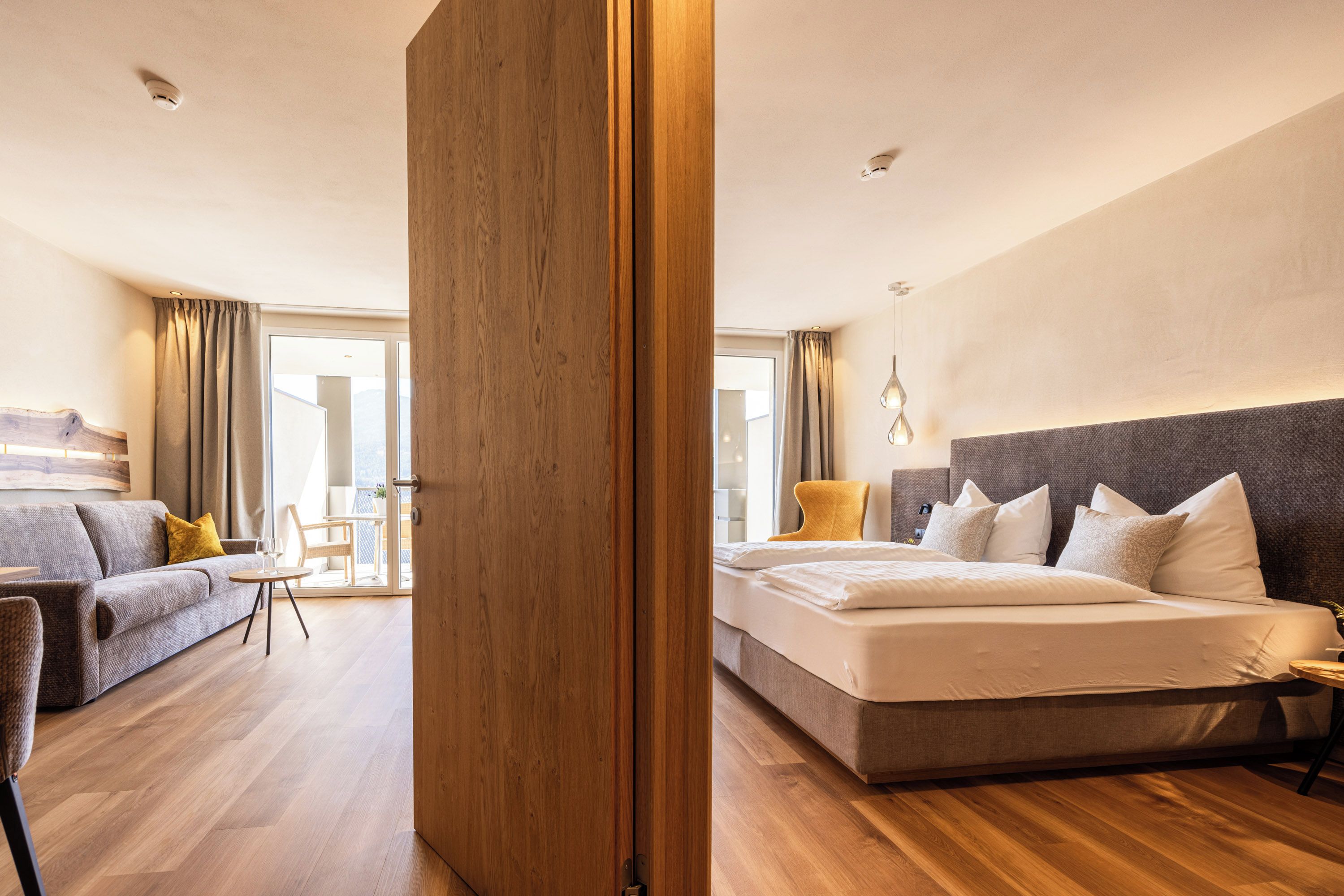 A hotel room with a wooden floor, a bed with white bedding, a grey sofa, and a dining area with a table and chairs.