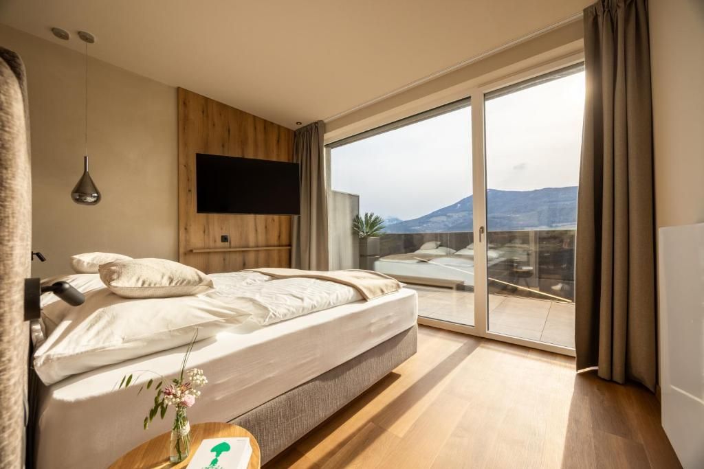 A bedroom with a bed, a TV mounted on the wall, and a large sliding glass door leading to a balcony with a view of mountains.