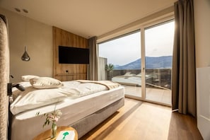 A bedroom with a bed, a TV mounted on the wall, and a large sliding glass door leading to a balcony with a view of mountains.