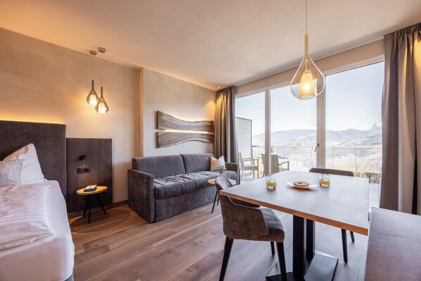 A hotel room with a bed, a sofa, a dining table, and a view of the mountains. - EGGER Apartments & Suites (Villanders)