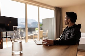 A person seated at a wooden table with a glass of water, a notebook, and a pen. In the background, there is a large window with a view of mountains and a modern interior with a chair and a table.