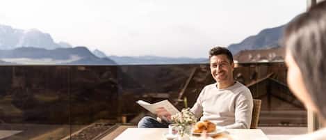 A person seated at a table with a view of mountains, a book, and a pastry.