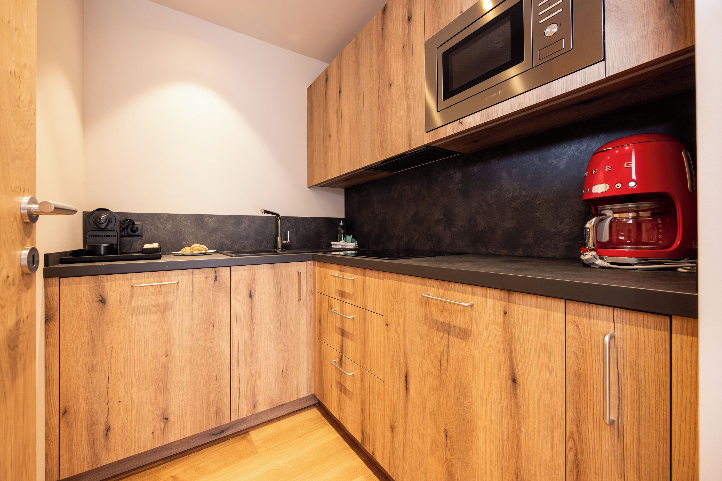 A modern kitchen with wooden cabinets, a black countertop, a stainless steel microwave, and a red coffee maker.